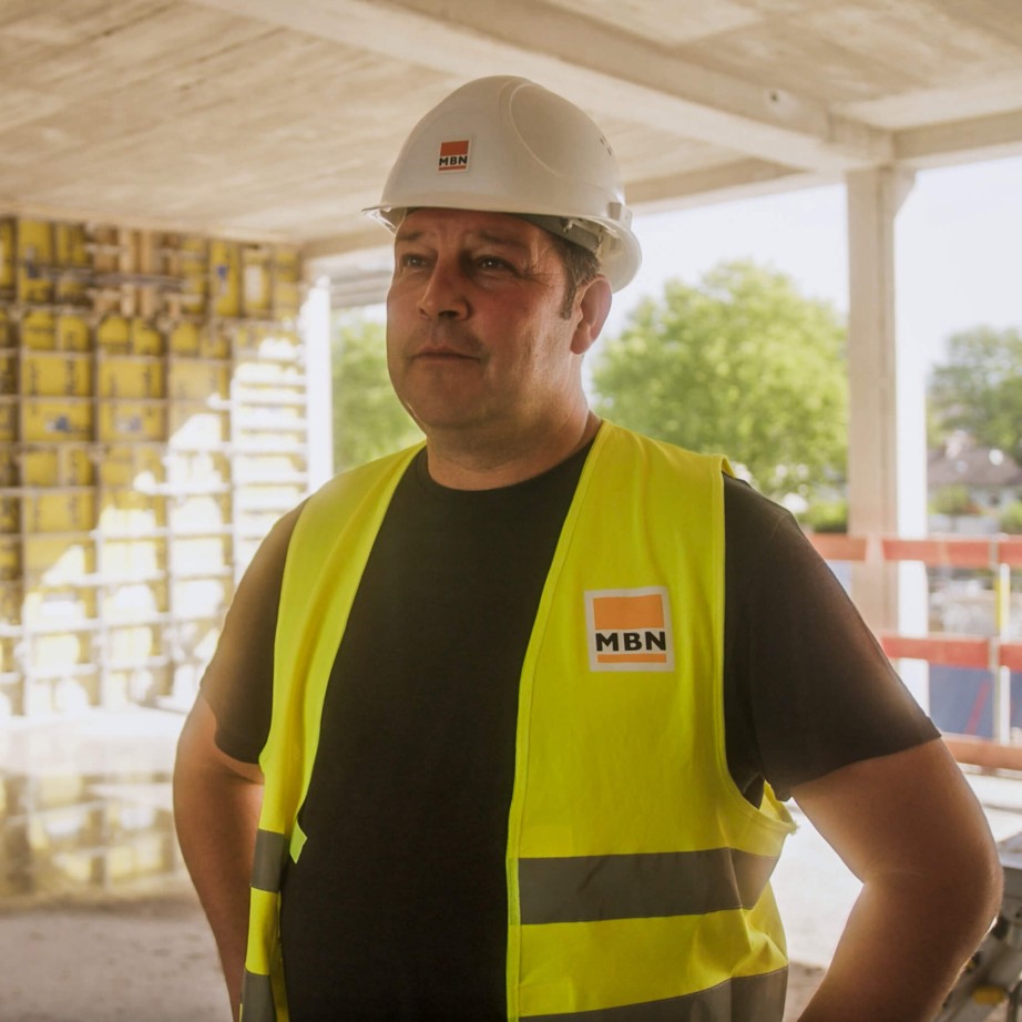 Polier der Firma MBN in Sicherheitsausrüstung auf der Baustelle mit der Bausoftware. Er steht mit dem Rücken zur offenen Fläche und der Rohbau seiner Baustelle ist erkennbar.