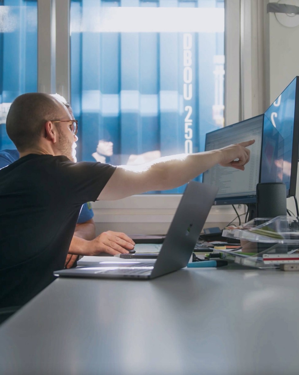 Zwei Personen bedienen am PC die intuitive Bausoftware für Bauleiter und Polier. Im Hintergrund ist ein Baucontainer zu sehen.